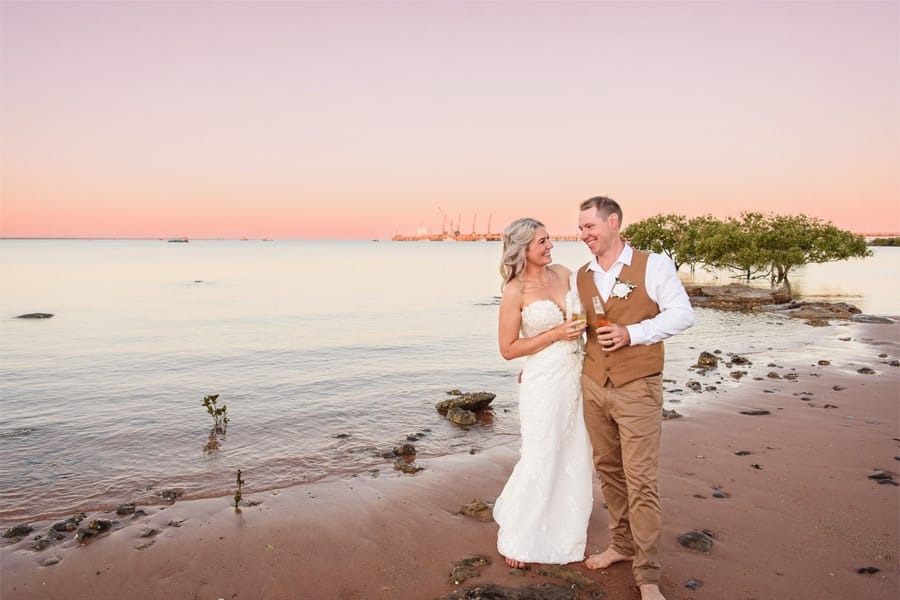 Bride and groom on the beach at sunset, holding drinks and smiling at each other.