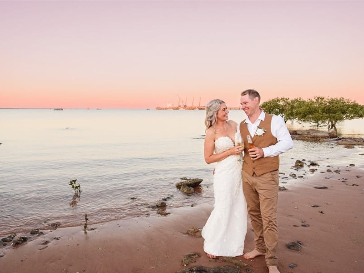 Bride and groom on the beach at sunset, holding drinks and smiling at each other.