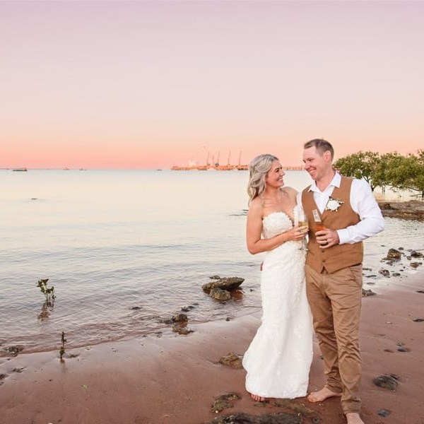 Bride and groom on the beach at sunset, holding drinks and smiling at each other.