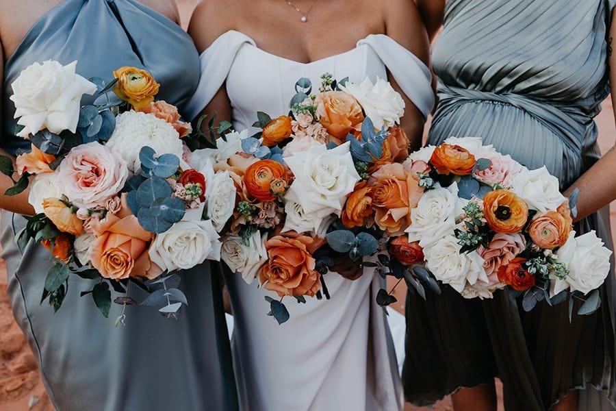 Three women in elegant dresses holding vibrant floral bouquets with orange, white, and pink flowers.