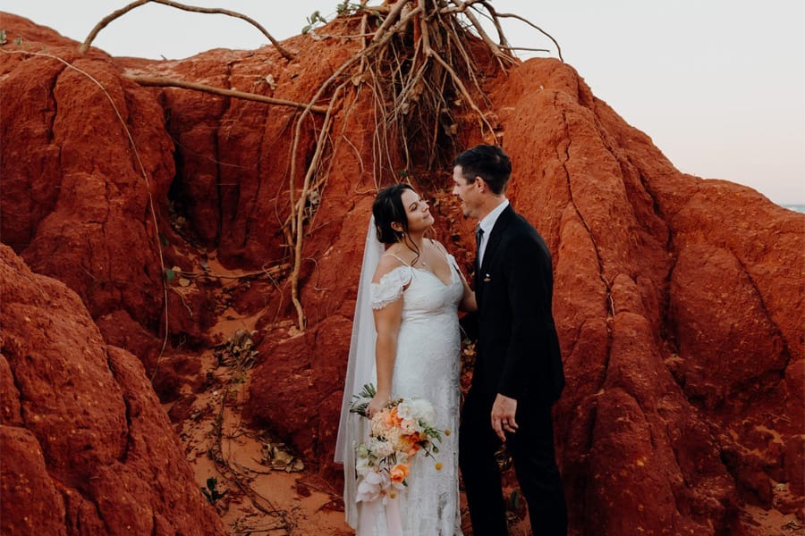 Bride and groom smiling at each other in red rocky landscape with exposed roots.