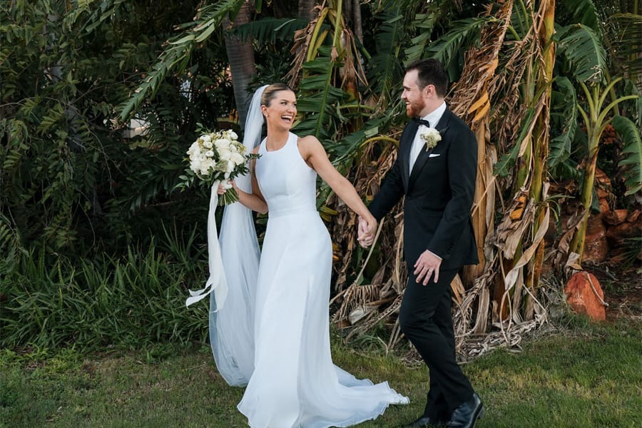 Bride and groom smiling, holding hands, walking outside with greenery in the background.