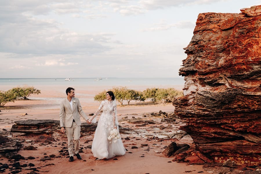Bride and groom walking on a rocky beach holding hands, large rock formation on the right.