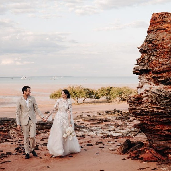 Bride and groom walking on a rocky beach holding hands, large rock formation on the right.