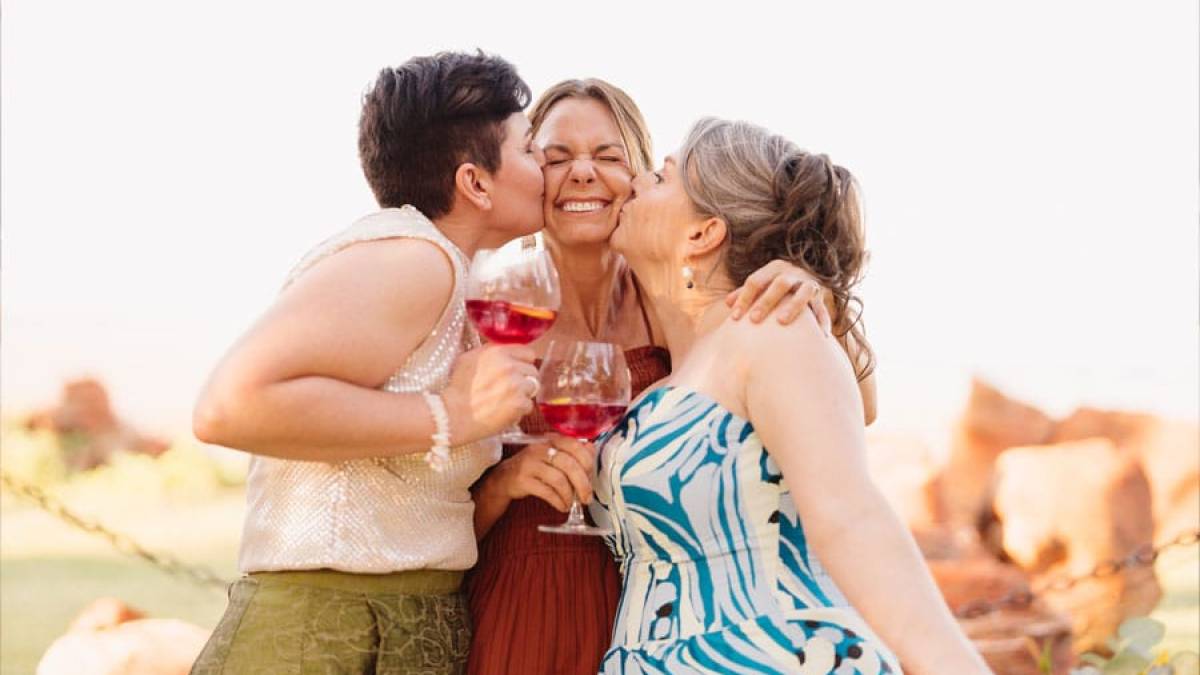 Three people smiling, holding drinks, two kissing one's cheeks outdoors.