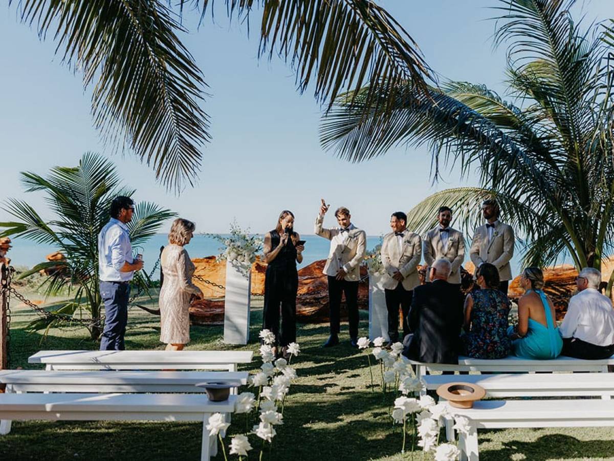 Outdoor wedding ceremony with guests seated under palm trees by the beach.