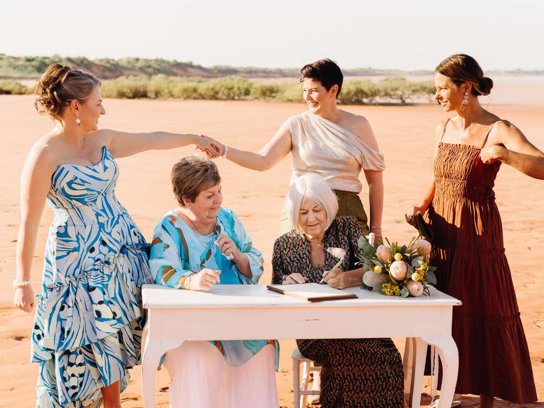 Five women celebrate around a table with a book and flowers on a beach.