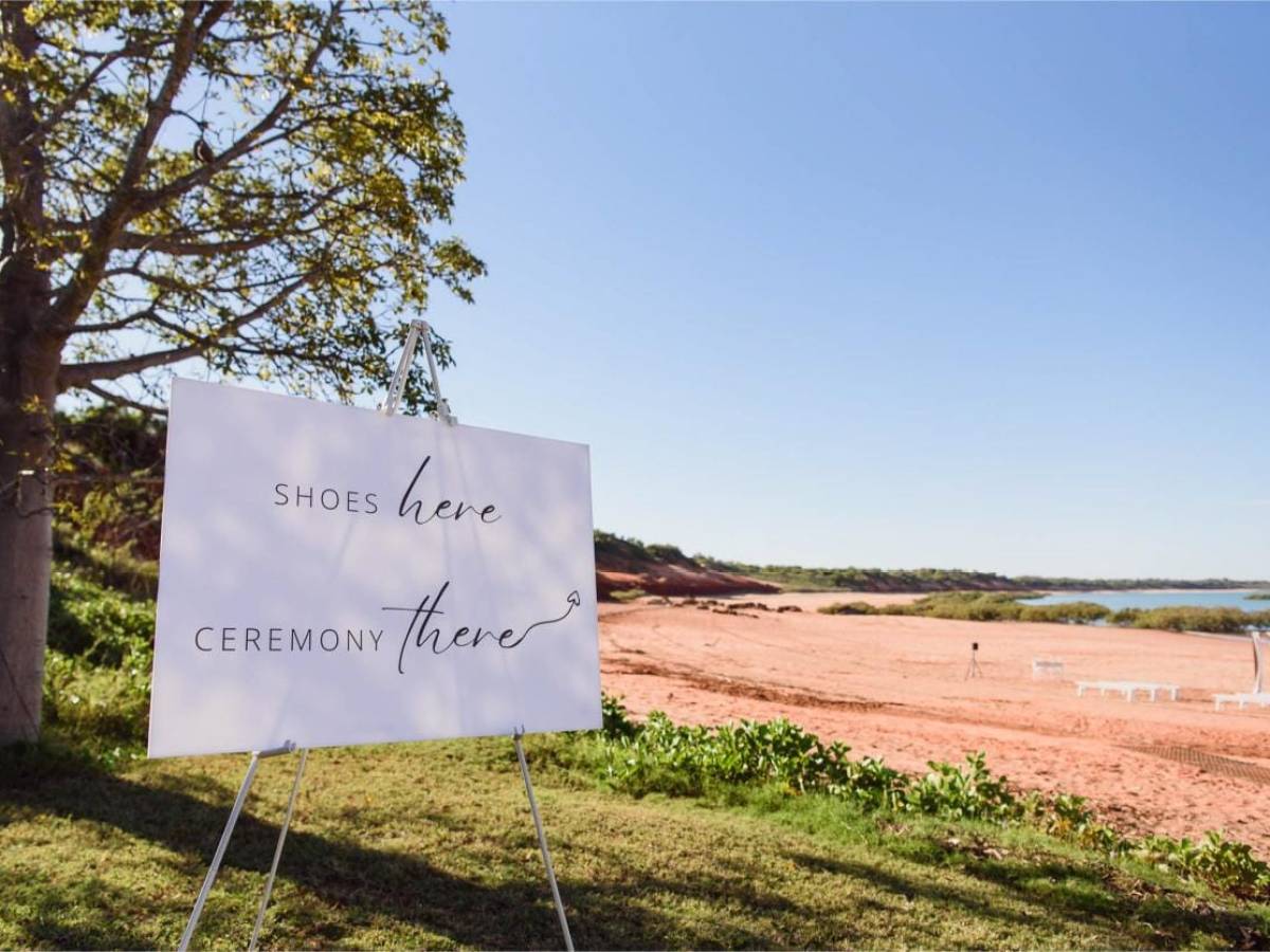 Beach with a sign reading 'Shoes here, ceremony there' next to a large tree.