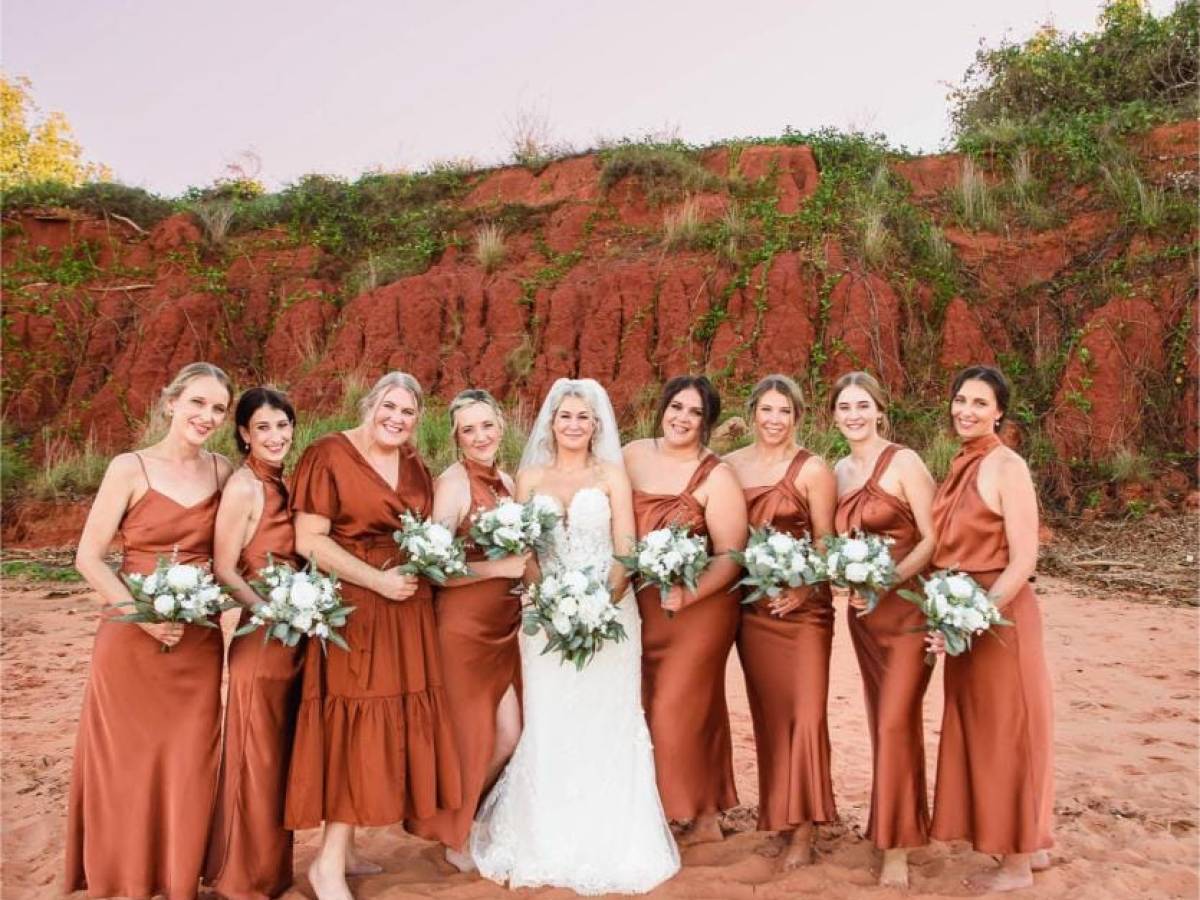 Bride with bridesmaids in orange dresses holding bouquets on a sandy terrain with a hillside background.