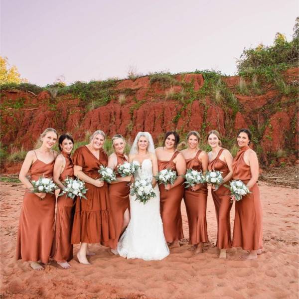 Bride with bridesmaids in orange dresses holding bouquets on a sandy terrain with a hillside background.