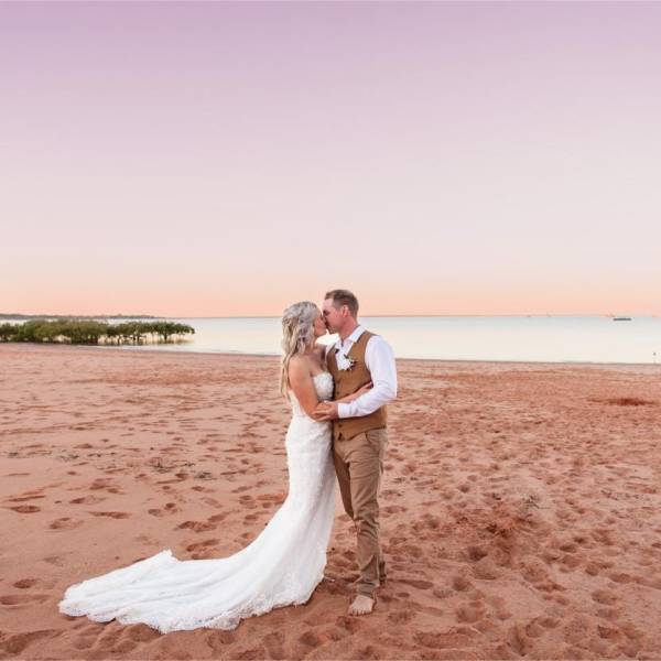 Couple in wedding attire kissing on a beach at sunset.