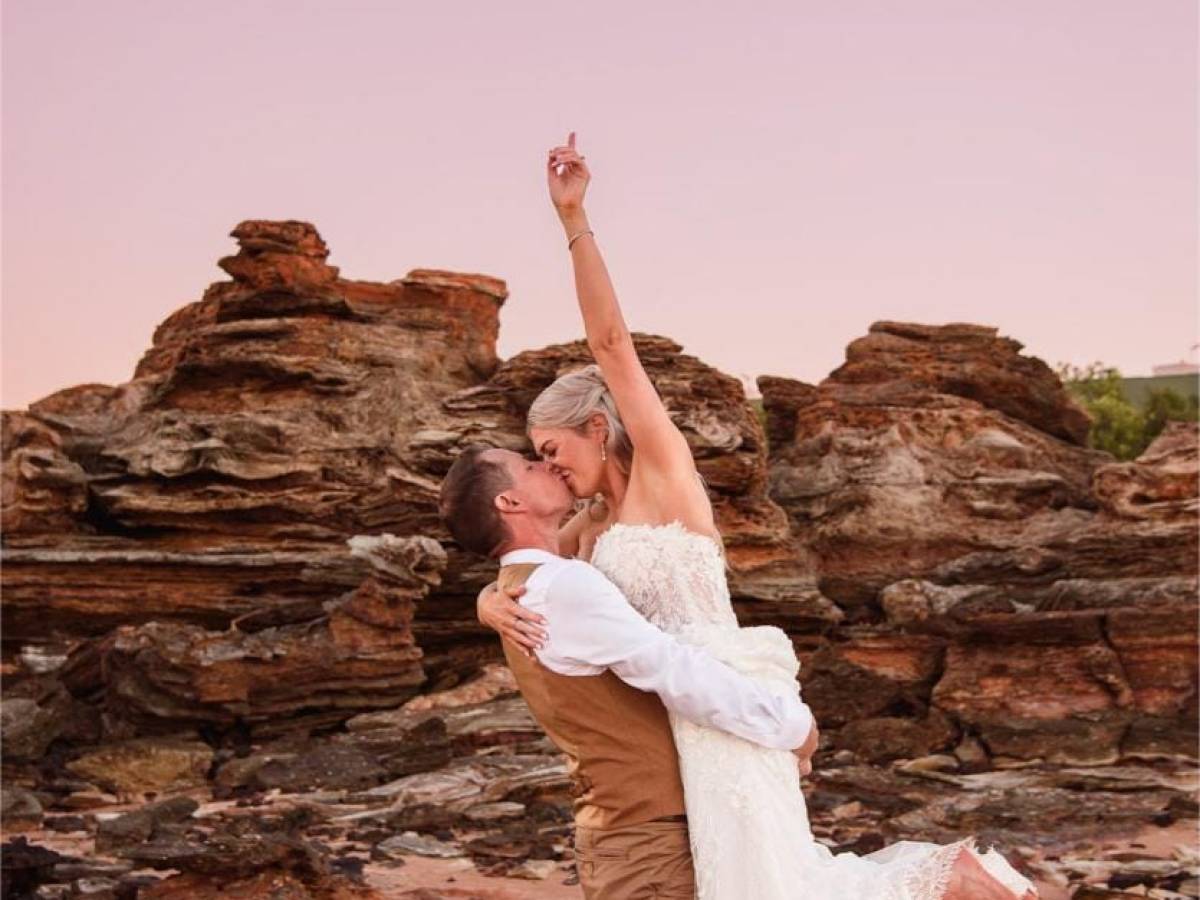 Bride and groom kiss on rocky beach at sunset, bride lifted in groom's arms, pink sky backdrop.