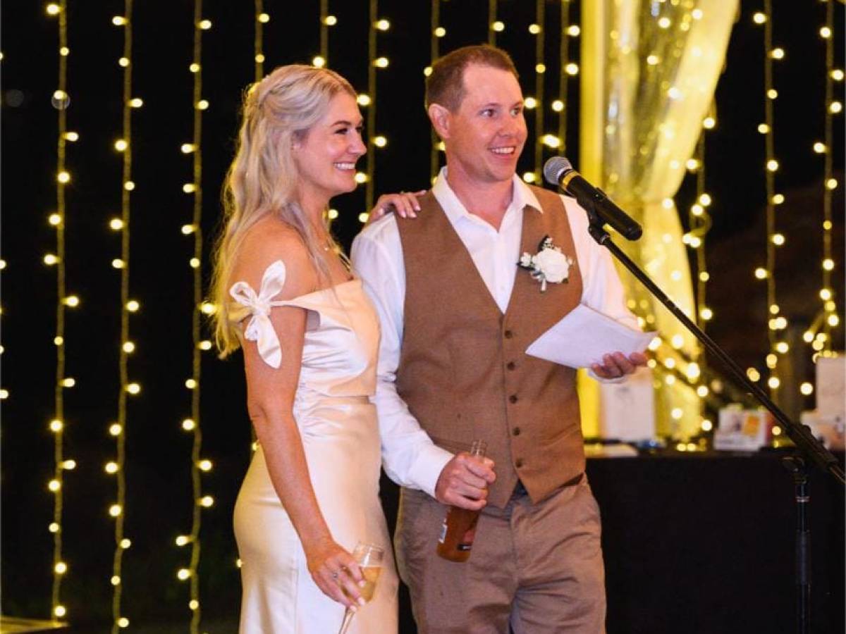 A couple stands under string lights, the man speaking into a microphone.