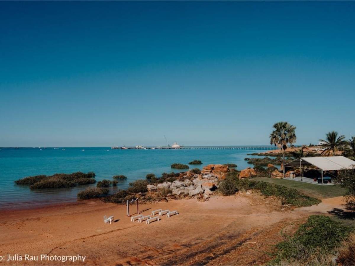 Beach with clear skies, scattered trees, a jetty in the distance, and a small shelter on the right.