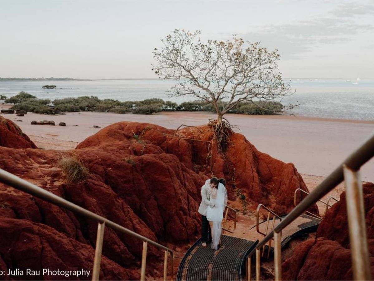 Couple embraces on staircase amid red rocks with beach and single tree in the background.