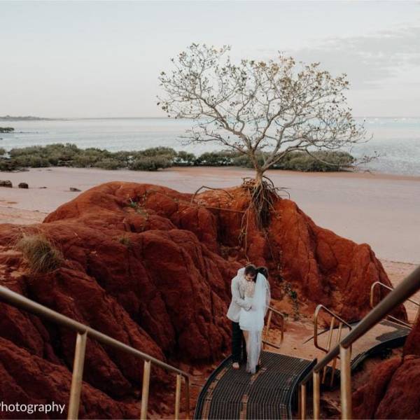 Couple embraces on staircase amid red rocks with beach and single tree in the background.