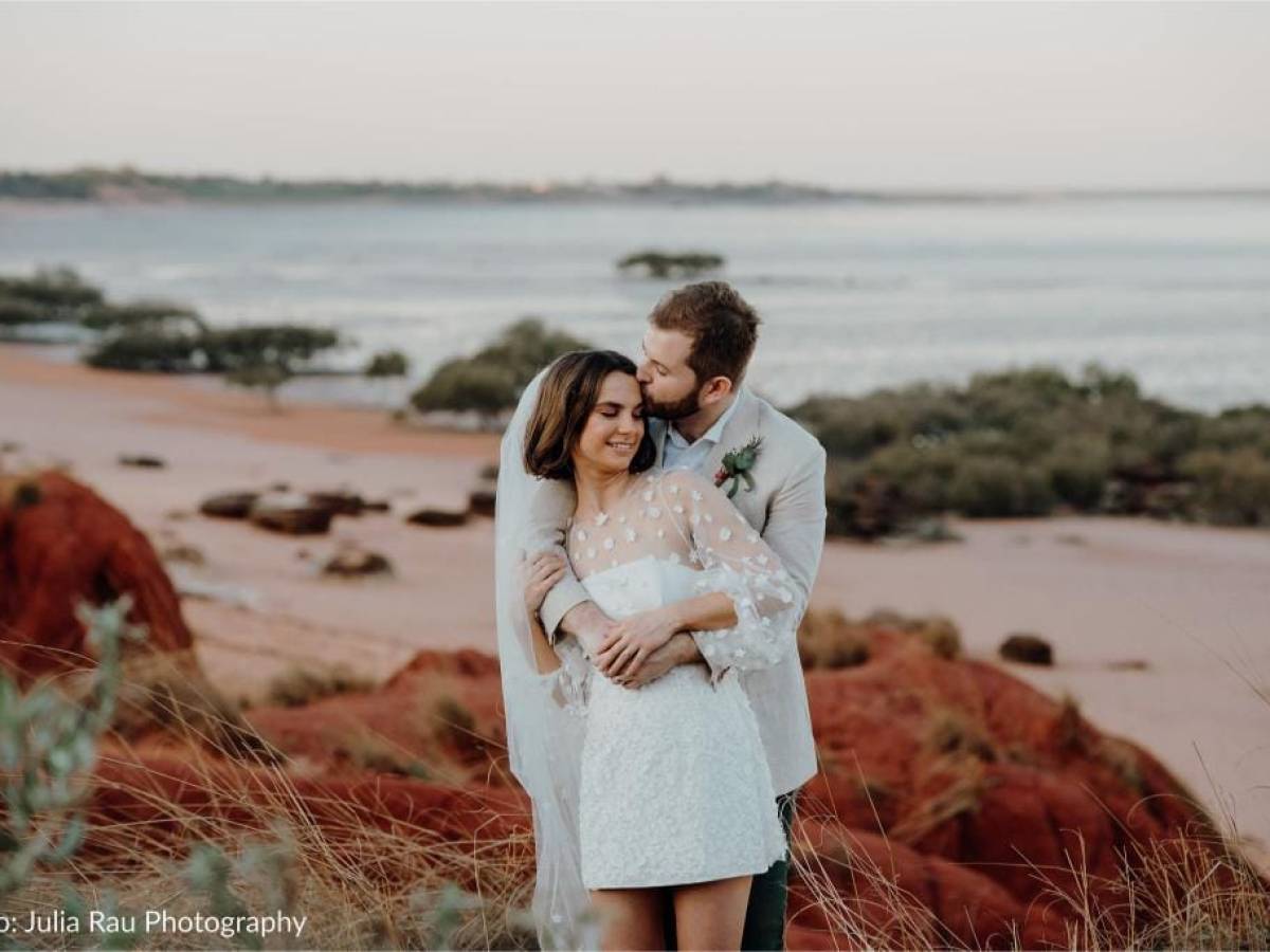 Couple embracing on a beach with red rocks, ocean in the background.