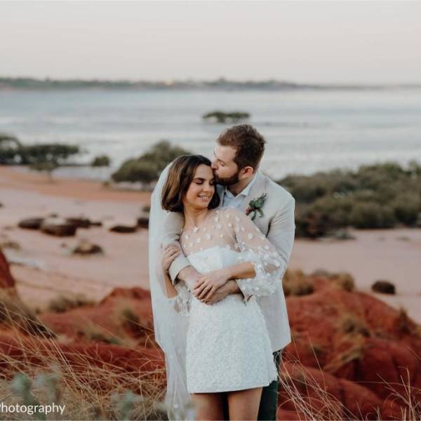 Couple embracing on a beach with red rocks, ocean in the background.