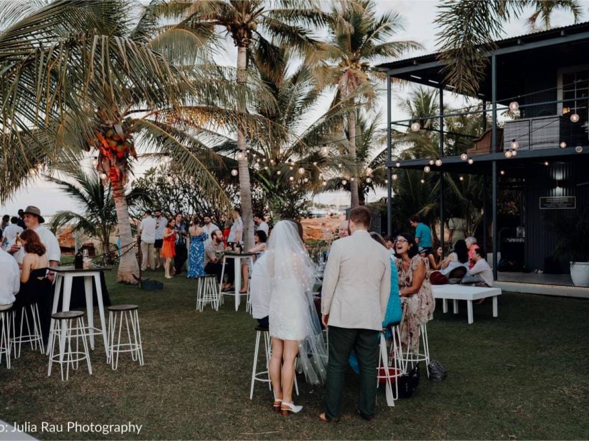Outdoor wedding reception with guests, palm trees, and string lights at sunset.