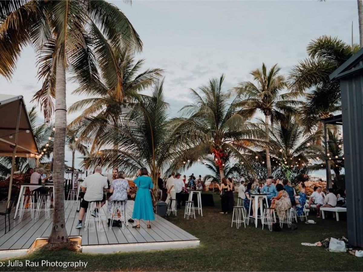 Outdoor party with people at tables, palm trees, and string lights at dusk.