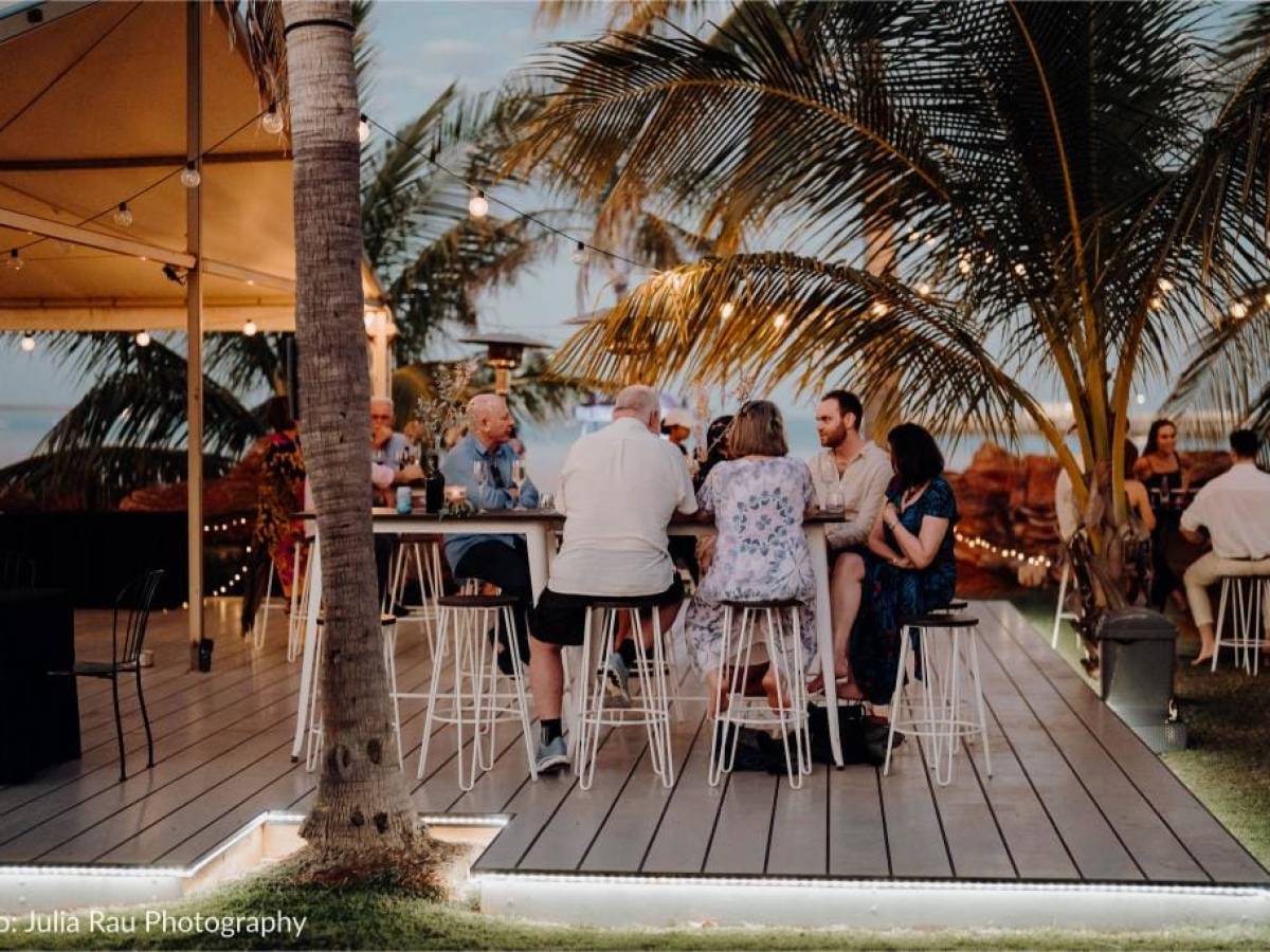 People sitting on a deck with string lights, palm trees, and ocean view at sunset.