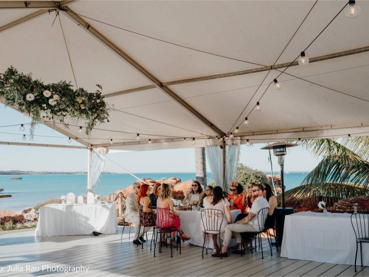 People seated at a table under a tent by the sea, with hanging lights and floral decoration.
