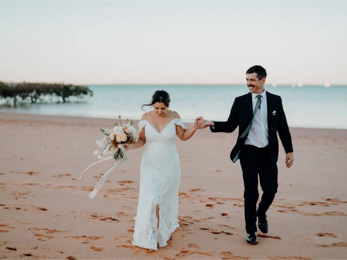 Bride and groom holding hands walking on a beach with ocean in background.