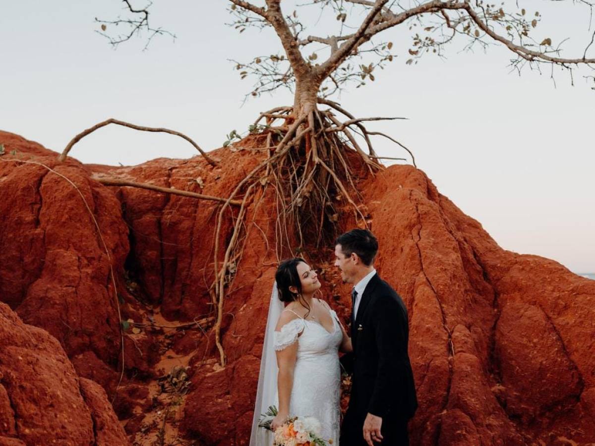 Bride and groom stand under a tree on red rock terrain.