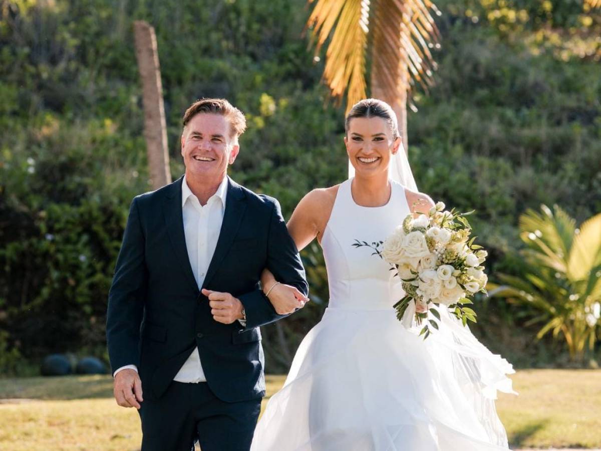 Bride in white dress holding bouquet, walking with man in dark suit on sunny day.