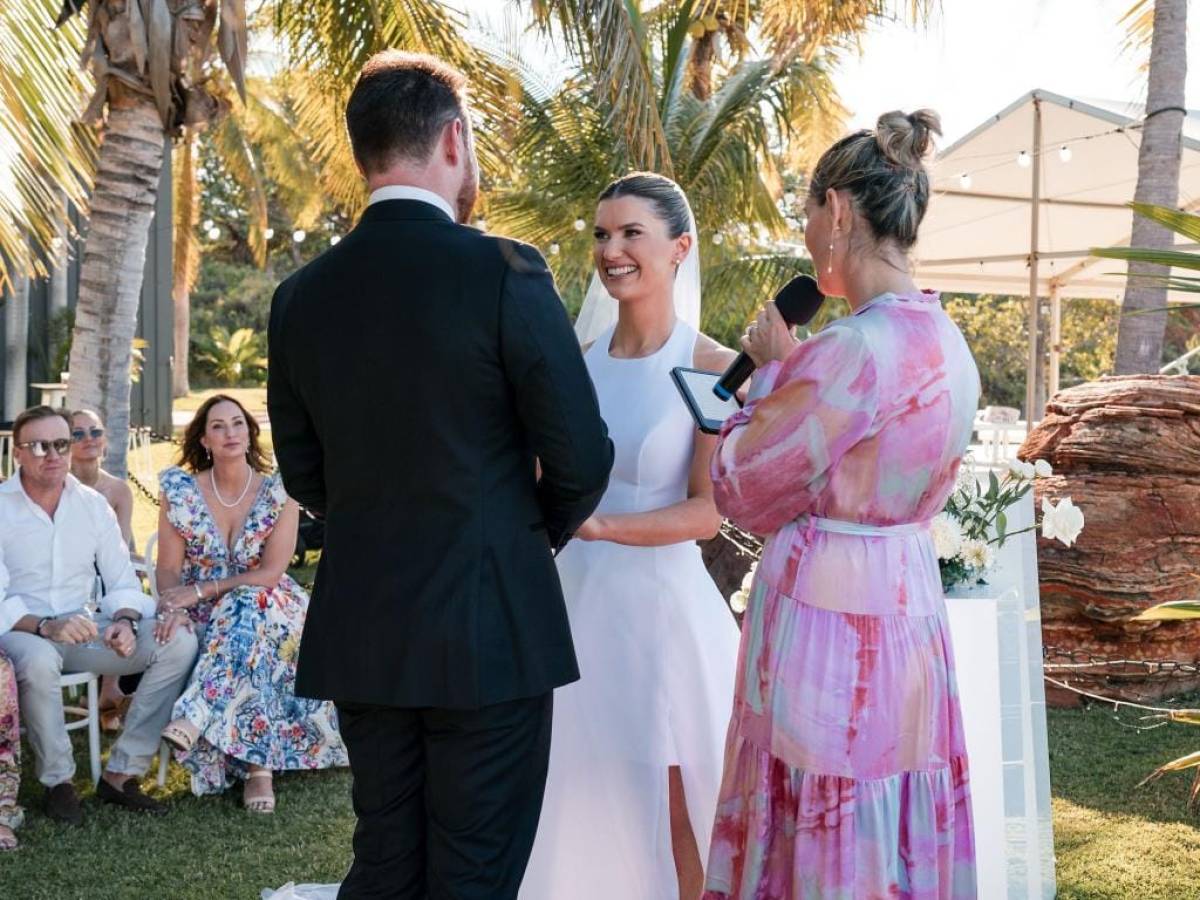 Outdoor wedding ceremony with a couple exchanging vows under palm trees, officiant holding microphone.