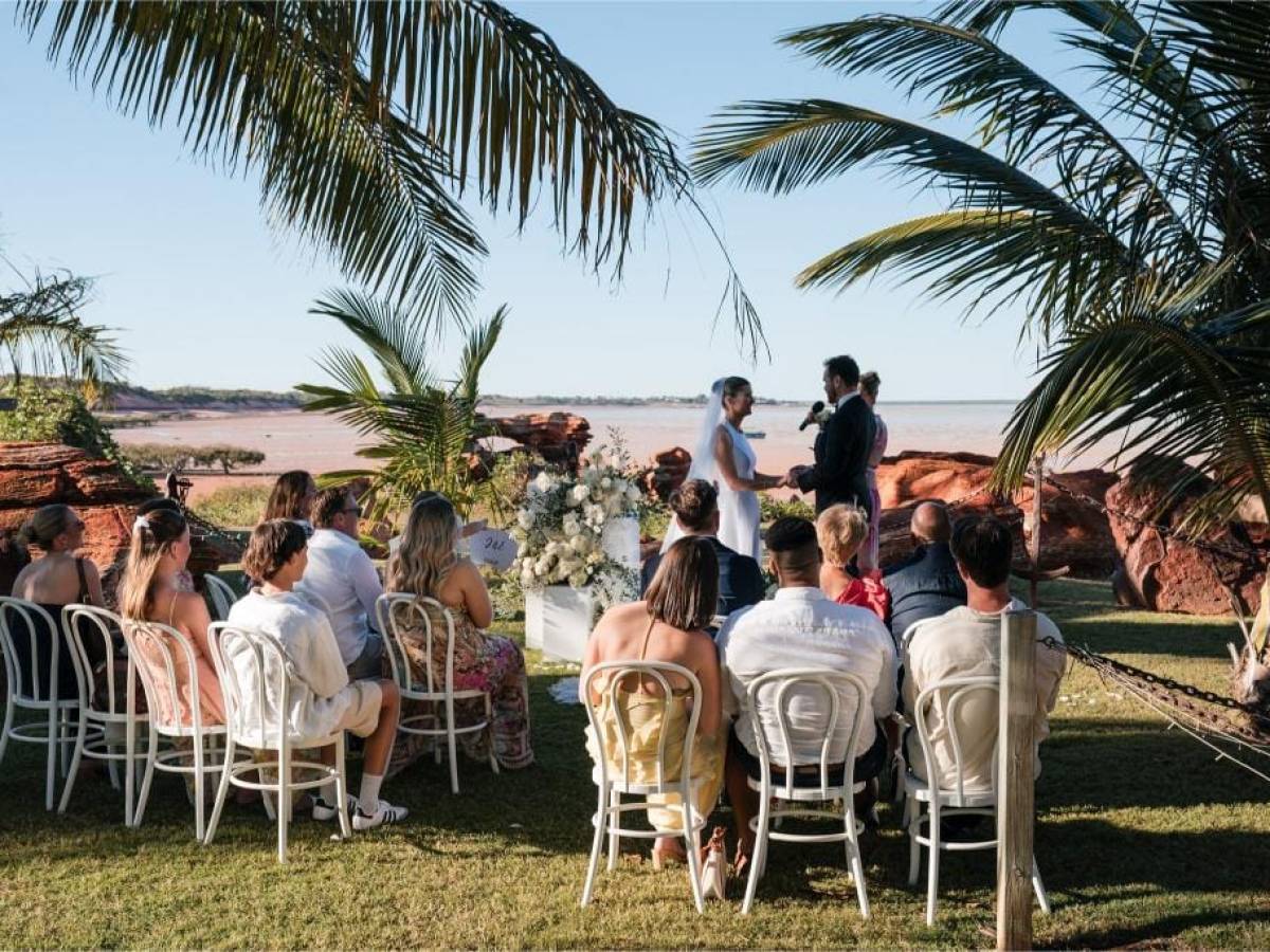 Outdoor beach wedding with guests seated, palm trees, and couple exchanging vows.