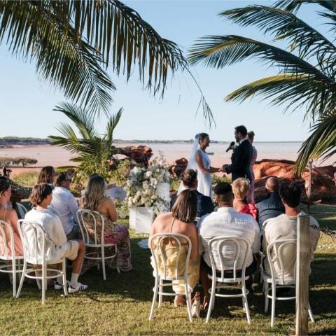 Outdoor beach wedding with guests seated, palm trees, and couple exchanging vows.