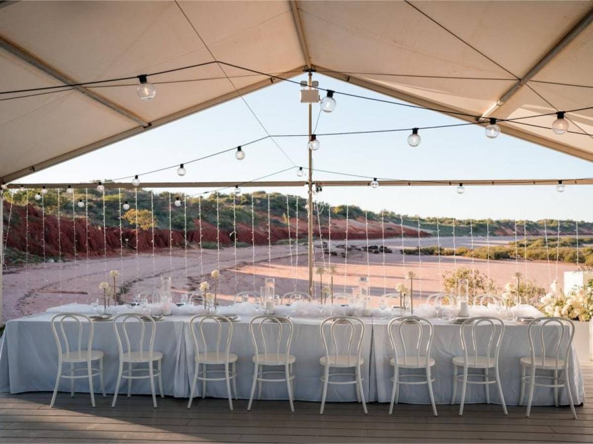 Elegant outdoor dining setup under tent with white chairs, tablecloth, and string lights.