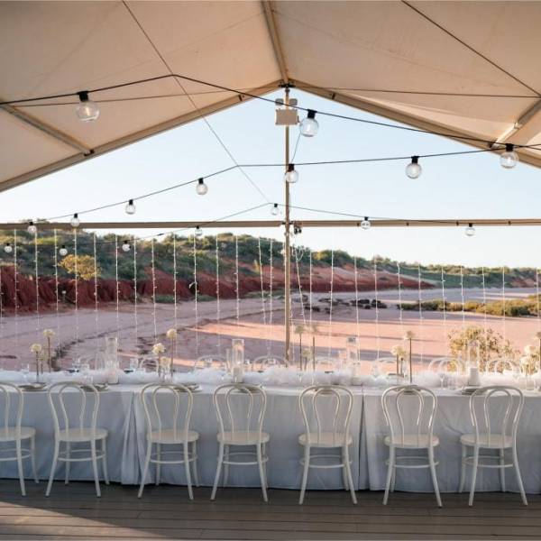 Elegant outdoor dining setup under tent with white chairs, tablecloth, and string lights.