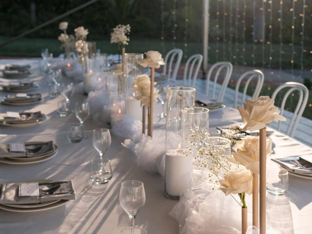 Elegant outdoor table setup with white roses, candles, plates, and glasses in the evening sun.
