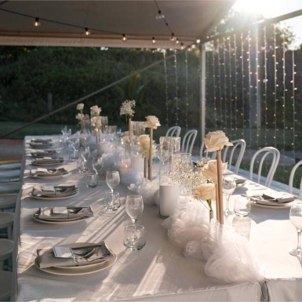 Elegant outdoor table setup with white tablecloth, chairs, floral centerpieces, and string lights.