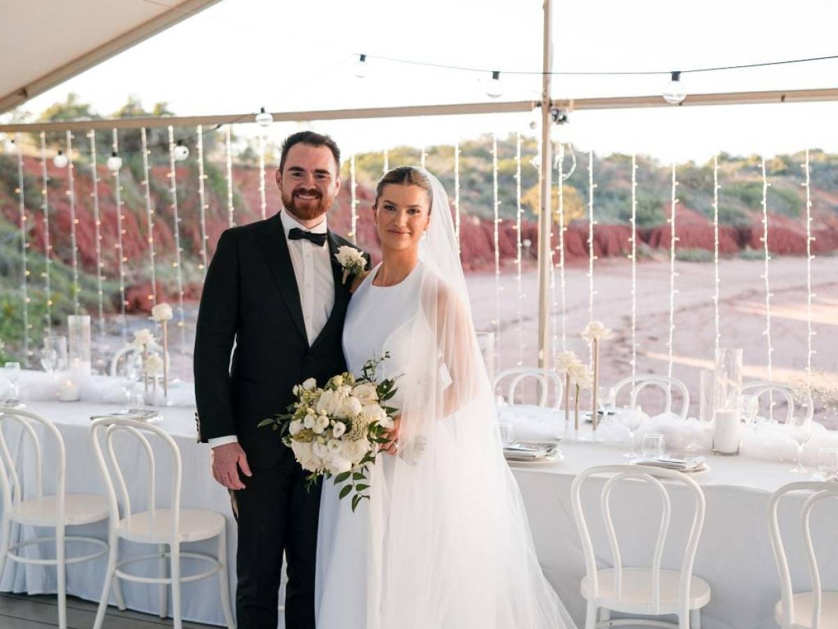 Bride and groom smiling under a tent with white decor and string lights, holding a bouquet.