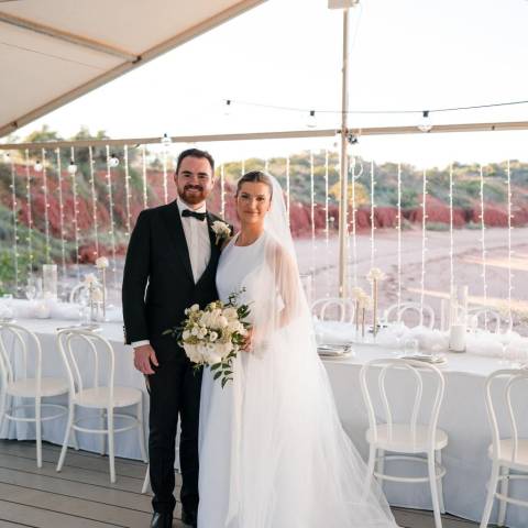 Bride and groom smiling under a tent with white decor and string lights, holding a bouquet.