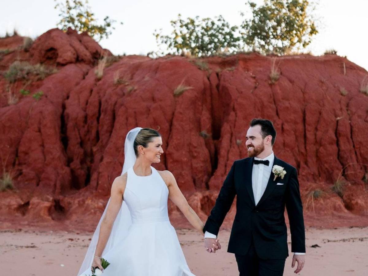 Bride and groom hold hands walking on sandy beach with red cliffs in background.
