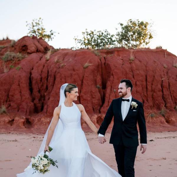Bride and groom hold hands walking on sandy beach with red cliffs in background.