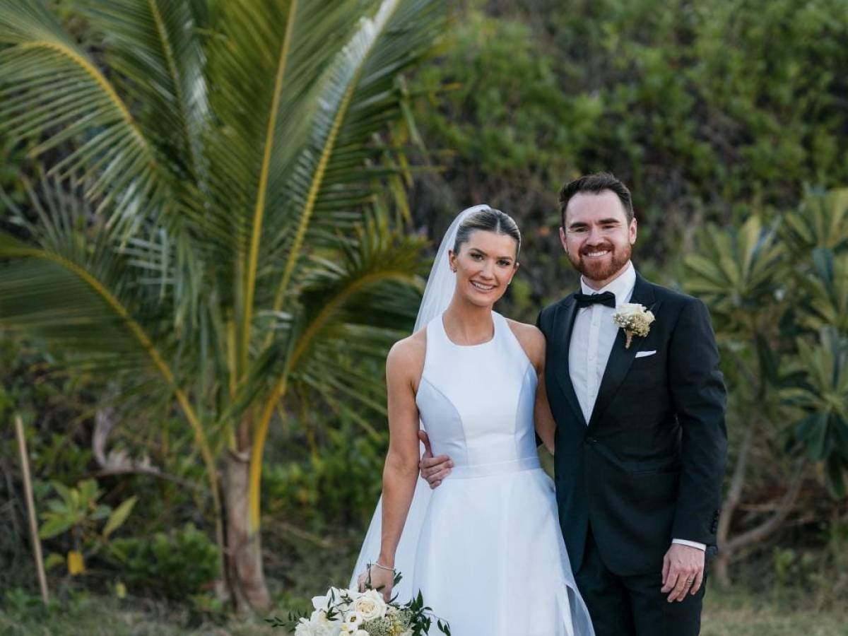 Bride in white dress and groom in black suit standing outdoors beside palm tree.