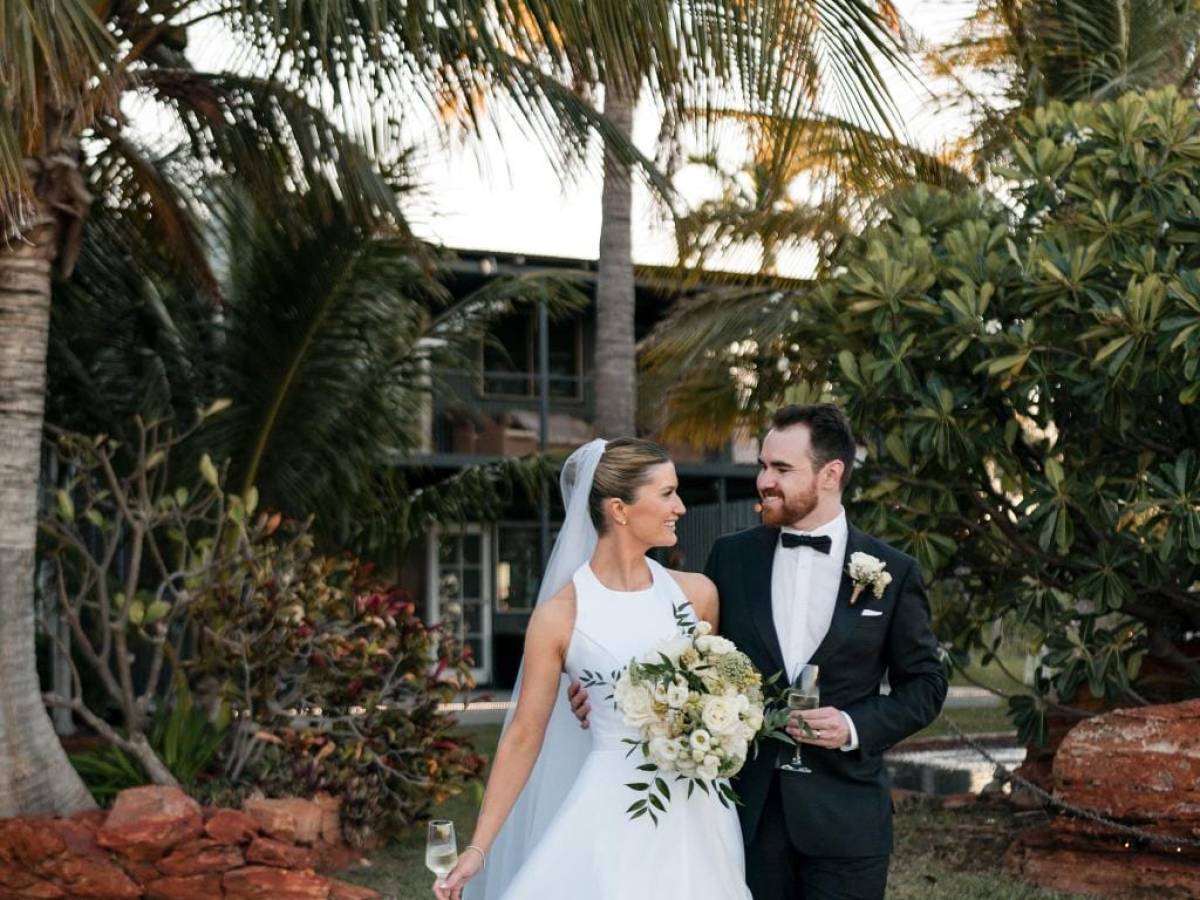Bride and groom smiling at each other outdoors, surrounded by palm trees.