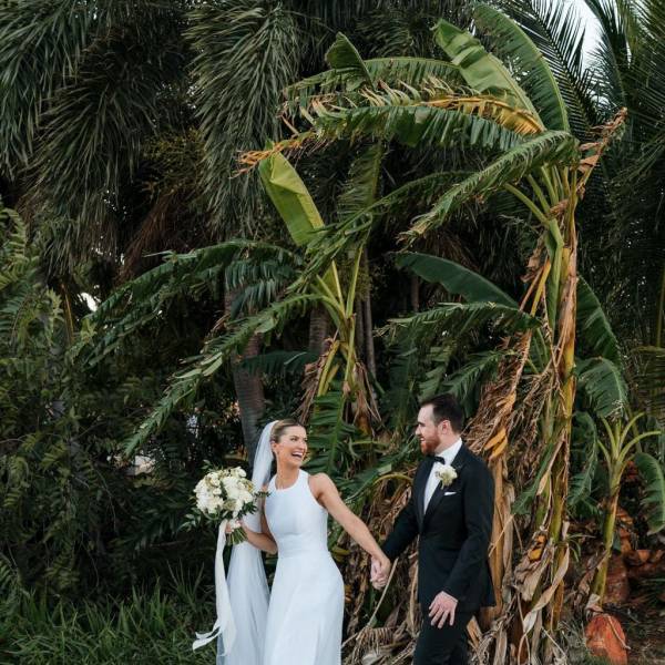 Bride and groom holding hands in front of lush tropical plants.