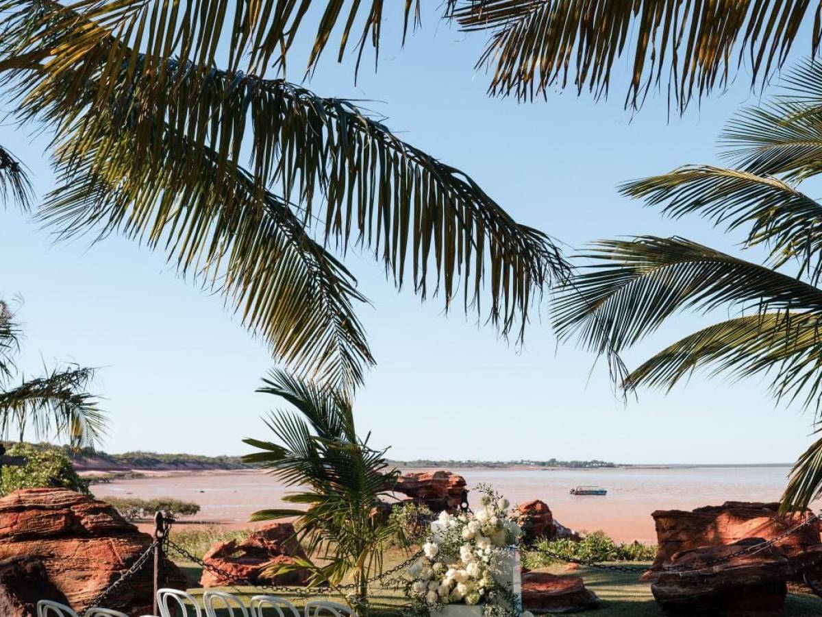 Outdoor wedding setup with white chairs, floral arrangements, and palm trees near a beach.