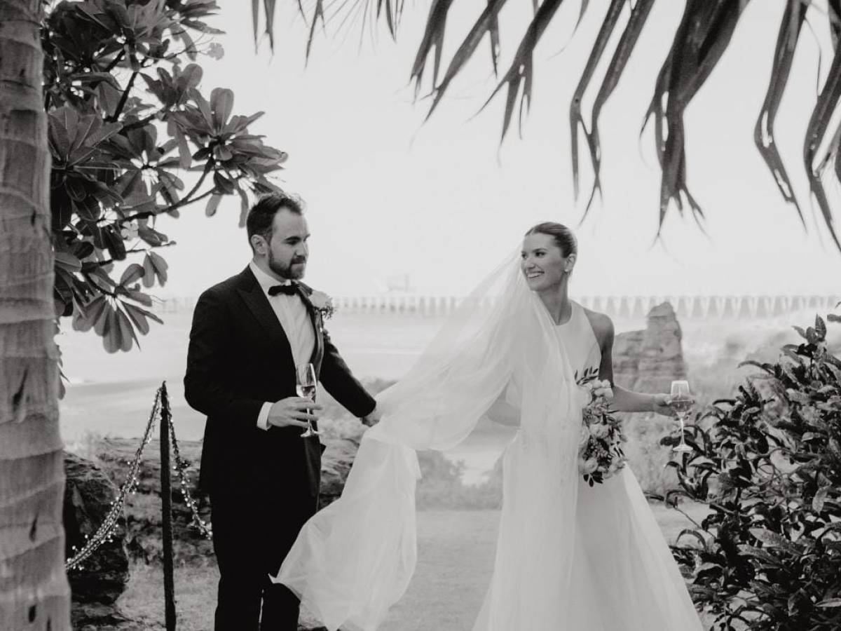 Black and white photo of a bride and groom holding hands outdoors under palm leaves.