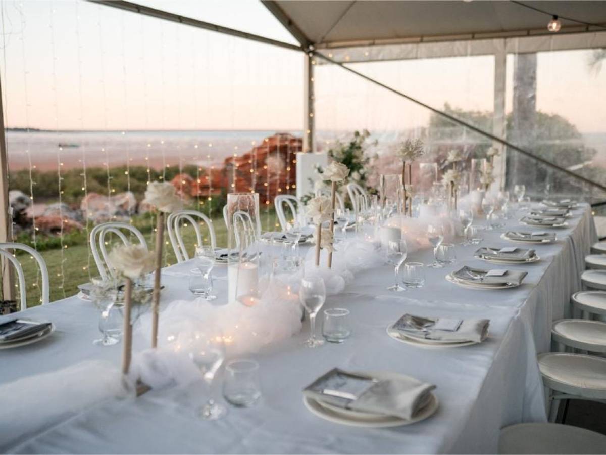 Elegant table setting in a tent with white chairs, floral arrangements, and soft lighting.