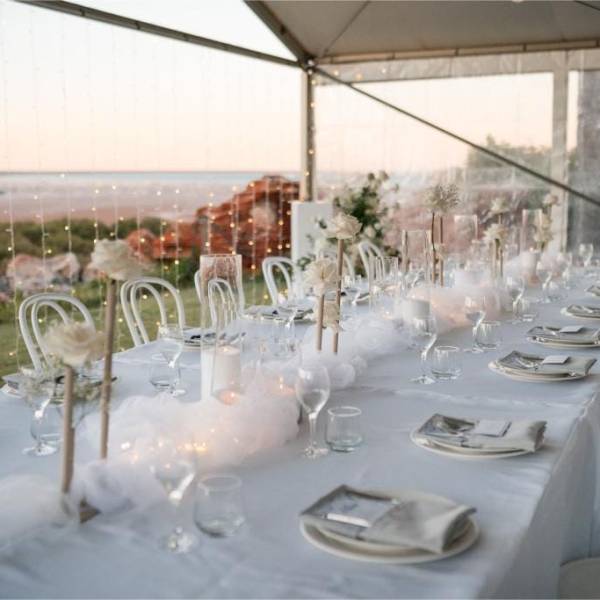 Elegant table setting in a tent with white chairs, floral arrangements, and soft lighting.