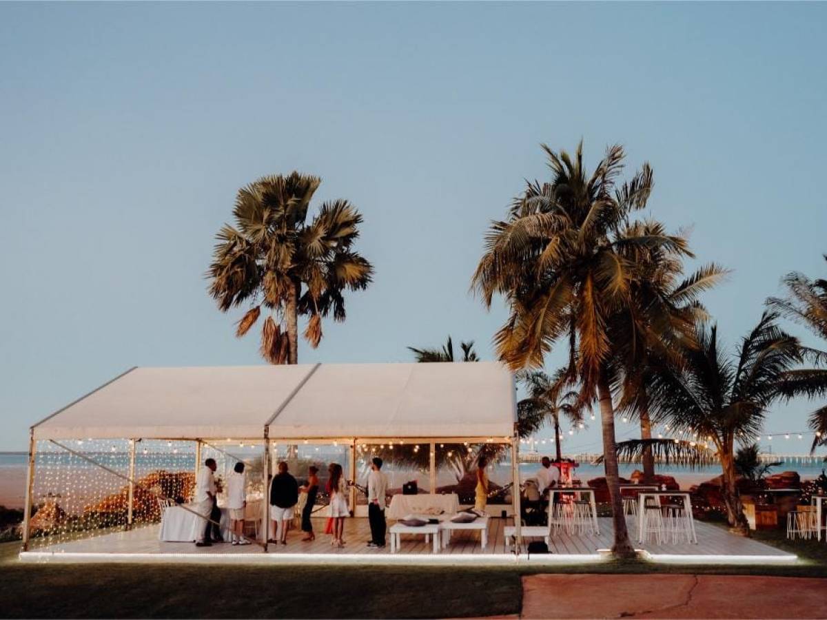 Outdoor party scene with people under a white tent, surrounded by palm trees and ocean view at sunset.