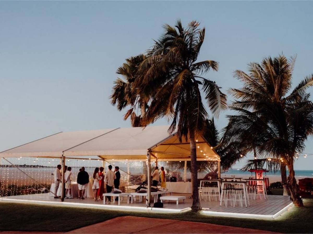 Outdoor event with people under a canopy near palm trees and ocean at dusk.
