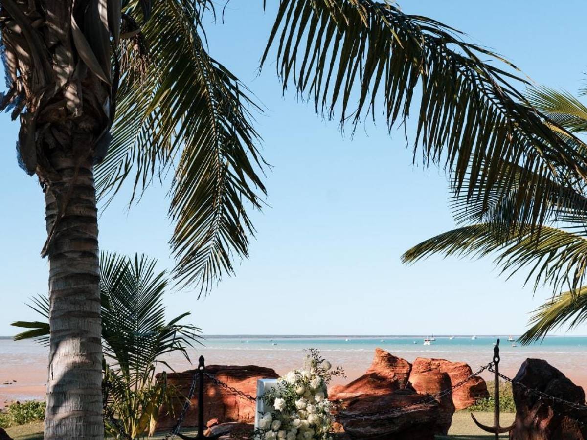 Outdoor beach wedding setup with white chairs, palm trees, and floral arrangement.